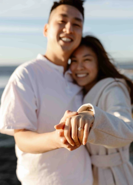 a photo of a man and woman, holding hands with engagement ring on her finger, wearing white, photoshoot near the sea